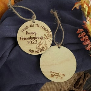 May include: Two round wooden ornaments with a twine string. The front of the ornament has the text "Friends are the family Happy Friendsgiving 2023 That you pick" with a pumpkin and floral design. The back of the ornament has the text "Chaimness Crafty Corner" with social media icons.