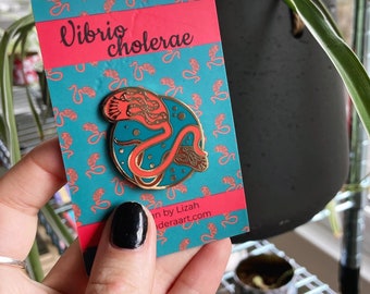 Puede incluir: Un pin de esmalte dorado con una ilustración estilizada de Vibrio cholerae, una bacteria. El pin está sobre un fondo turquesa con un patrón repetido de la bacteria. La tarjeta dice "Vibrio cholerae" en rojo.