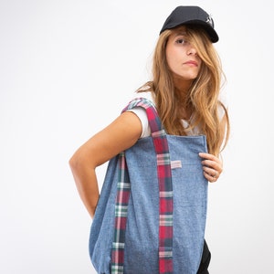 Op de afbeelding: Een vrouw draagt een zwarte baseballpet met een wit T-shirt en een zwarte broek en houdt een blauwe draagtas vast met rood-groene geruite banden.