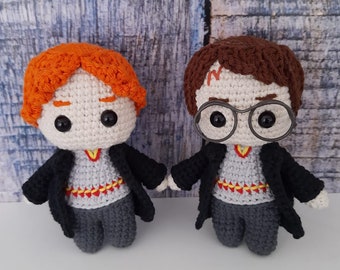 May include: Two crocheted Harry Potter characters, one with red hair and the other with brown hair, both wearing black robes and grey pants. The character with red hair has a scar on his forehead.