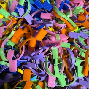 以下が含まれることがあります： カラフルなプラスチック製のカップホルダーの山。ホルダーはオレンジ、グリーン、ピンク、パープル、ブルー、イエローなど、さまざまな色合いです。ホルダーはすべて、上部に円形の開口部のあるY字型をしています。