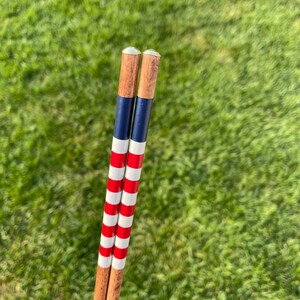 May include: Two wooden croquet mallets with a patriotic design. The mallets feature alternating red and white stripes, and blue and white stripes. The mallets have a wooden shaft and a brass-colored top.