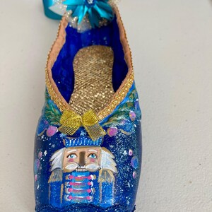 May include: A blue ballet slipper ornament decorated with a painted Nutcracker face, gold glitter, and a blue and white flower.
