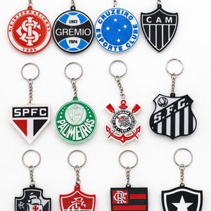 Pode incluir: Uma cole&ccedil;&atilde;o de chaveiros com v&aacute;rios logotipos de equipas. Os chaveiros t&ecirc;m diferentes formas, incluindo c&iacute;rculos e escudos, e apresentam nomes e logotipos de equipas em vermelho, azul, verde, preto e branco. Cada chaveiro tem uma corrente e um anel prateados.