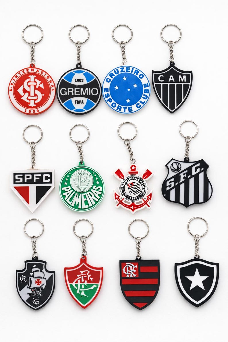 Pode incluir: Uma cole&ccedil;&atilde;o de chaveiros com v&aacute;rios logotipos de equipas. Os chaveiros t&ecirc;m diferentes formas, incluindo c&iacute;rculos e escudos, e apresentam nomes e logotipos de equipas em vermelho, azul, verde, preto e branco. Cada chaveiro tem uma corrente e um anel prateados.