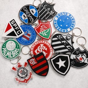 Pode incluir: Uma cole&ccedil;&atilde;o de chaveiros coloridos com v&aacute;rios logotipos e emblemas de times de futebol. Os chaveiros t&ecirc;m v&aacute;rias formas, incluindo escudos e c&iacute;rculos, e apresentam nomes e logotipos de times em branco, vermelho, preto e verde. Cada chaveiro tem um chaveiro prateado.