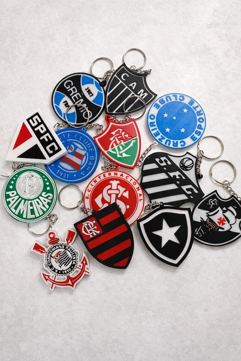 Pode incluir: Uma cole&ccedil;&atilde;o de chaveiros coloridos com v&aacute;rios logotipos e emblemas de times de futebol. Os chaveiros t&ecirc;m v&aacute;rias formas, incluindo escudos e c&iacute;rculos, e apresentam nomes e logotipos de times em branco, vermelho, preto e verde. Cada chaveiro tem um chaveiro prateado.