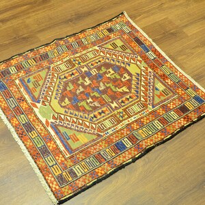 May include: A small, square rug with a geometric design. The rug features a central medallion with a repeating pattern in shades of brown, red, blue, and green. The border has a repeating pattern of rectangles and zigzags. The rug is placed on a wooden floor.
