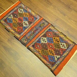 May include: A rectangular, hand-woven rug with a geometric pattern. The rug features a central design of colorful squares in red, blue, yellow, and green, framed by a border of orange and other colors. The rug is placed on a wooden floor.