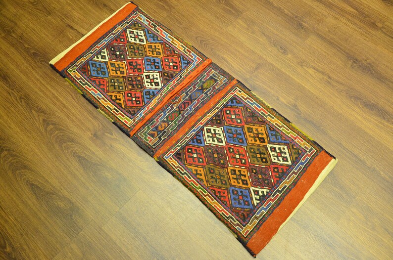 May include: A rectangular, hand-woven rug with a geometric pattern. The rug features a central design of colorful squares in red, blue, yellow, and green, framed by a border of orange and other colors. The rug is placed on a wooden floor.