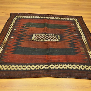 Puede incluir: Una alfombra cuadrada tejida a mano con un diseño geométrico. La alfombra presenta un campo central rojo oscuro con dos rayas negras y un cuadrado central con un diseño. El borde es un patrón repetitivo en oro y marrón.