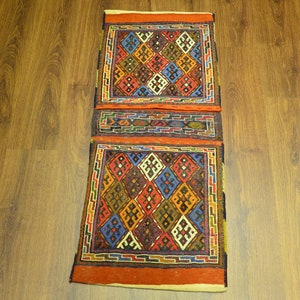 May include: A rectangular, hand-woven rug with a geometric pattern. The rug features a repeating diamond design in shades of red, blue, yellow, and brown, framed by a border of orange and green. The rug is placed on a wooden floor.
