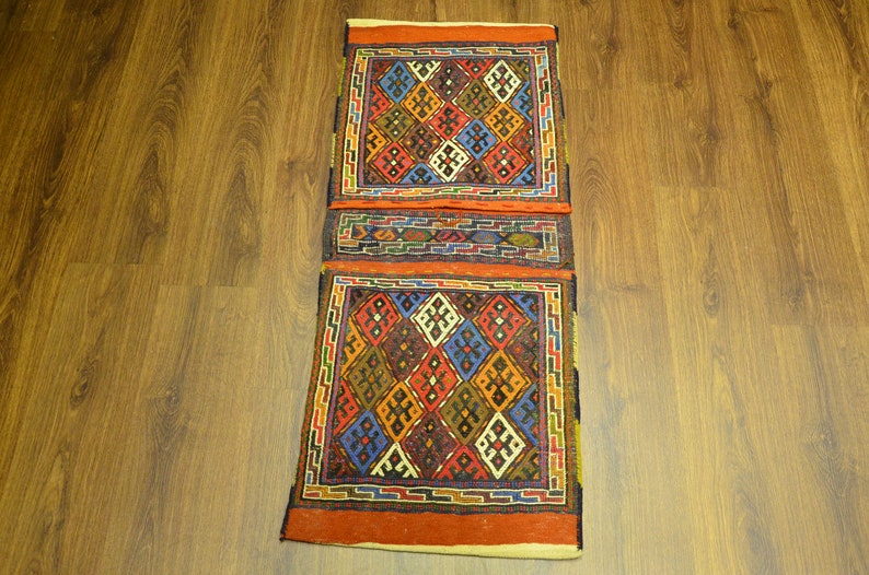 May include: A rectangular, hand-woven rug with a geometric pattern. The rug features a repeating diamond design in shades of red, blue, yellow, and brown, framed by a border of orange and green. The rug is placed on a wooden floor.