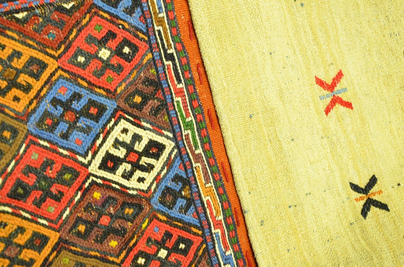 May include: Close-up of a section of a woven rug with a geometric pattern. The rug features a vibrant mix of colors, including red, blue, brown, and orange, arranged in a repeating diamond design. The other section is a light yellow color with red and black cross-like designs.