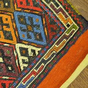 May include: Close-up of a hand-woven rug with a geometric pattern. The rug features a variety of colors, including orange, red, blue, and black. The design includes diamond and square shapes, with intricate detailing.