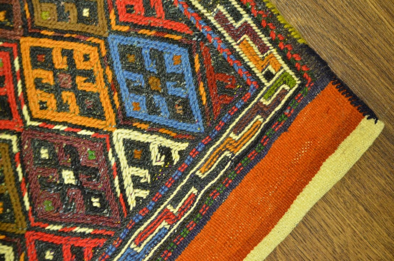 May include: Close-up of a hand-woven rug with a geometric pattern. The rug features a variety of colors, including orange, red, blue, and black. The design includes diamond and square shapes, with intricate detailing.
