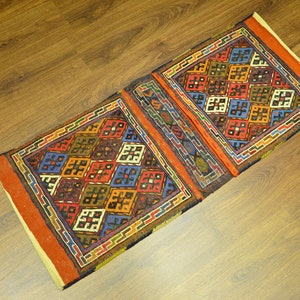 May include: A rectangular, patterned rug with a geometric design. The rug features a repeating pattern of colorful squares in shades of red, blue, orange, and green. The rug has a brown border and is placed on a wooden surface.