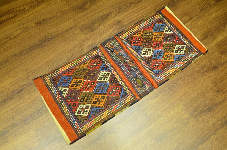 May include: A rectangular, patterned rug with a geometric design. The rug features a repeating pattern of colorful squares in shades of red, blue, orange, and green. The rug has a brown border and is placed on a wooden surface.