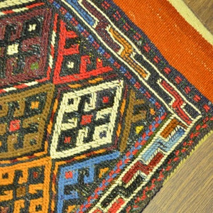 May include: Close-up of a section of a colorful, hand-woven rug. The rug features a geometric pattern with squares in shades of red, blue, brown, and orange. The design includes intricate cross-like motifs and borders.