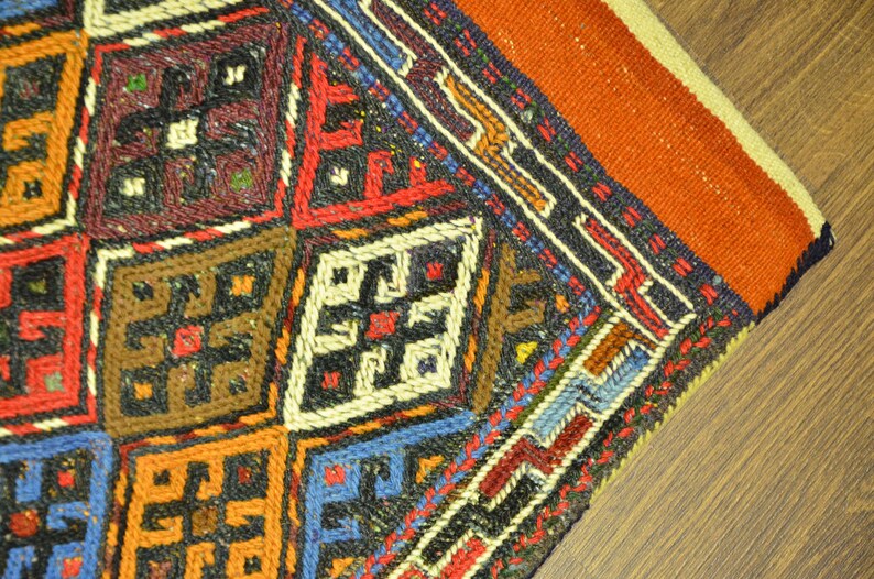 May include: Close-up of a section of a colorful, hand-woven rug. The rug features a geometric pattern with squares in shades of red, blue, brown, and orange. The design includes intricate cross-like motifs and borders.