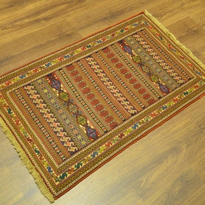 Puede incluir: Alfombra rectangular tejida a mano con un detallado patrón geométrico. La alfombra presenta rayas verticales en rojo, naranja y marrón, con toques de azul, verde y amarillo. El borde es marrón oscuro con un patrón repetido. La alfombra tiene flecos.