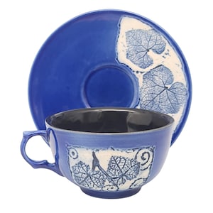 May include: A blue ceramic teacup and saucer set. The cup and saucer are a vibrant blue with a white leaf pattern. The interior of the cup is black. The saucer is round and the cup has a handle.