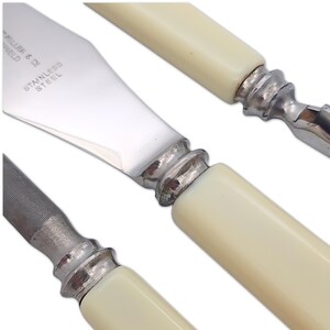 May include: Close-up of stainless steel knives with off-white handles. The blades are shiny and reflect light. The handles have a classic design with a silver metal bolster. The text "STAINLESS STEEL" is visible on the blade.