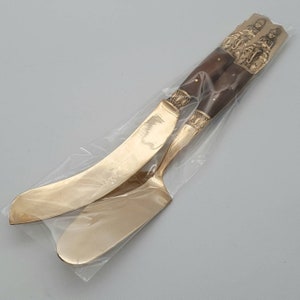 May include: A set of two gold-toned butter knives with ornate handles. The handles are made of dark wood and feature intricate carvings of figures and animals. The knives are packaged in a clear plastic bag.