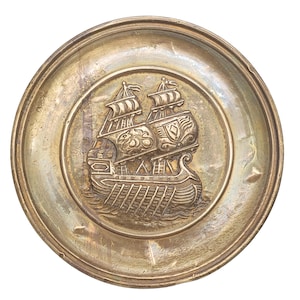 May include: A round, brass tray with a raised, detailed image of a ship with sails. The ship is in the center of the tray and is surrounded by a smooth, brass surface.