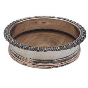 May include: A silver-plated drinks coaster with a decorative beaded edge and a copper-coloured band. The coaster has a wooden interior and is designed to hold a glass or cup. The coaster is round and has a classic, elegant design.