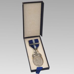 May include: A silver medal with a blue and silver ribbon, presented in a black box. The medal has an oval design with two figures and a star-shaped top. The box is lined with a beige fabric.