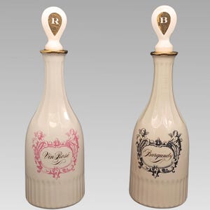 May include: Two off-white ceramic decanters with matching stoppers. One features a pink label with the text Vin Rosé and a letter R on the stopper. The other features a black label with the text Burgundy and a letter B on the stopper.