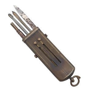 May include: A vintage brass multi-tool case featuring a rectangular body with a sliding mechanism. Three metal implements, including a pointed blade with visible oxidation, a thin rod, and a hooked tool, extend from the top. A small ring is attached to the base.