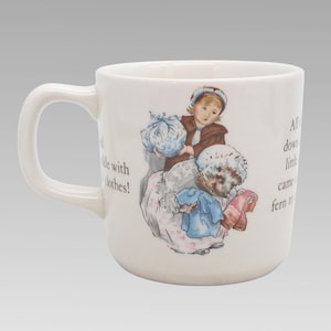 May include: A white ceramic mug featuring a colourful illustration of a child and a hedgehog carrying bundles of clothes. The design includes printed text on both sides of the central image, depicting a scene with soft pastel colours and detailed line work.