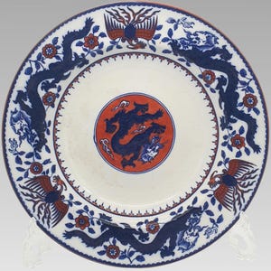 May include: A white ceramic plate with a red and blue design. The plate features a central red circle with a blue dragon, surrounded by blue dragons, phoenixes, and floral patterns. The rim has a red border.