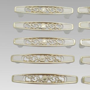 May include: A collection of five elongated drawer pulls arranged in a vertical column. Each pull features a metallic gold frame surrounding a white centre panel with an intricate, openwork scroll design and a central circular accent piece.