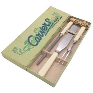 May include: A vintage carving set in a green box with the word "Carvers" in blue and green script. The set includes a carving knife, a sharpening steel, a carving fork, and a spatula, all with cream-colored handles. The box reads "Made in Sheffield England."
