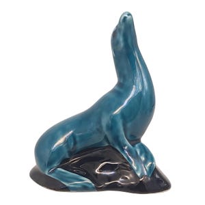 May include: A teal glazed ceramic seal figurine. The seal is depicted with its head and neck raised, sitting on a dark blue base. This decorative figurine is a charming addition to any home.