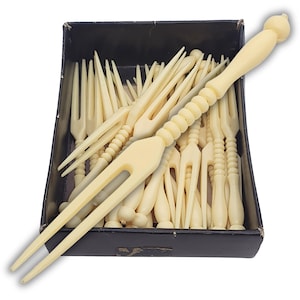May include: A collection of antique cream-coloured cocktail forks in a black box. The forks have a two-pronged design with decorative handles. The box is filled with many forks, with one fork in the foreground. The handles have a ribbed design.