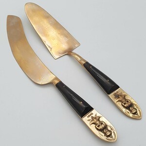 May include: A set of two gold-toned cake servers with black handles. The handles are decorated with intricate carvings of a mythical creature. The servers are made of metal and have a polished finish.