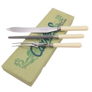 May include: A vintage carving set with a knife, fork, and sharpening steel, each with cream-colored handles and silver accents. The set rests on a light green box with a decorative design and the text "Made in England".