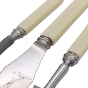 May include: Close-up of antique silverware with ivory-colored handles and stainless steel blades. The blades are stamped with the words "STAINLESS STEEL" and "MILLER & CO SHEFFIELD". The silverware includes a knife, a spreader, and a serrated knife.