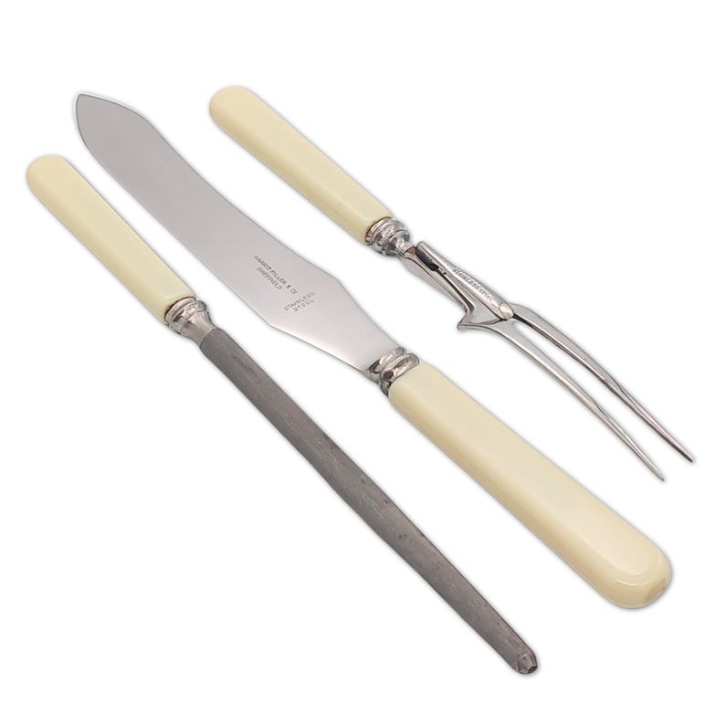 May include: A set of antique silverware, including a knife, a fork, and a sharpening steel. The knife and fork have ivory-colored handles and silver accents. The sharpening steel is a dark gray color.