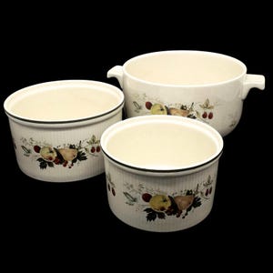 May include: Three off-white ceramic bowls with a fruit and leaf design. The bowls are different sizes, with a black rim and a cream-colored interior. The largest bowl has handles on either side.