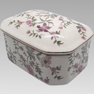 May include: A rectangular ceramic box with a lid, featuring a floral pattern in shades of pink, purple, and green on a cream background. The box has a brown trim and a glossy finish, suitable for storing jewelry or small items.