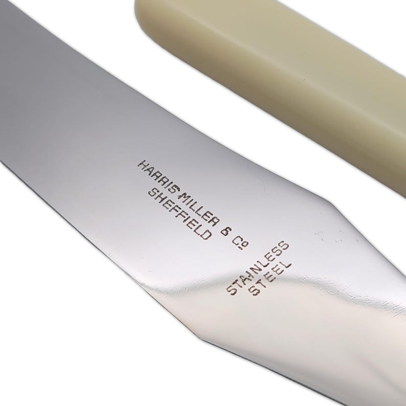 May include: Close-up of a stainless steel knife blade with a cream-colored handle. The blade is engraved with "HARRIS MILLER & CO SHEFFIELD STAINLESS STEEL". The knife has a pointed tip and a smooth, reflective surface.