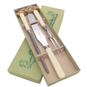 May include: A vintage carving set in a green box. The set includes a carving knife, fork, and sharpening steel, all with cream-colored handles and silver accents. The box is labeled "Made in Sheffield England".