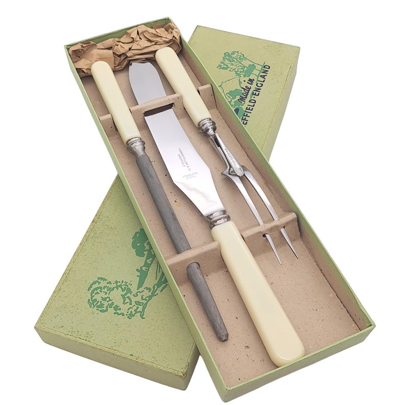May include: A vintage carving set in a green box. The set includes a carving knife, fork, and sharpening steel, all with cream-colored handles and silver accents. The box is labeled "Made in Sheffield England".