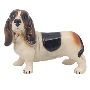 May include: A ceramic Basset Hound figurine. The dog has a white body with black, brown, and tan markings. The figurine has long, floppy ears and a sad expression. The dog is in a standing position.