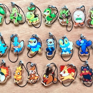 Op de afbeelding: Een verzameling kleurrijke sleutelhangers met stripfiguren. De sleutelhangers tonen verschillende wezens in tinten groen, blauw, oranje en rood. Elke sleutelhanger heeft een zwart koord voor bevestiging en wordt gepresenteerd op een lichtbruin oppervlak.